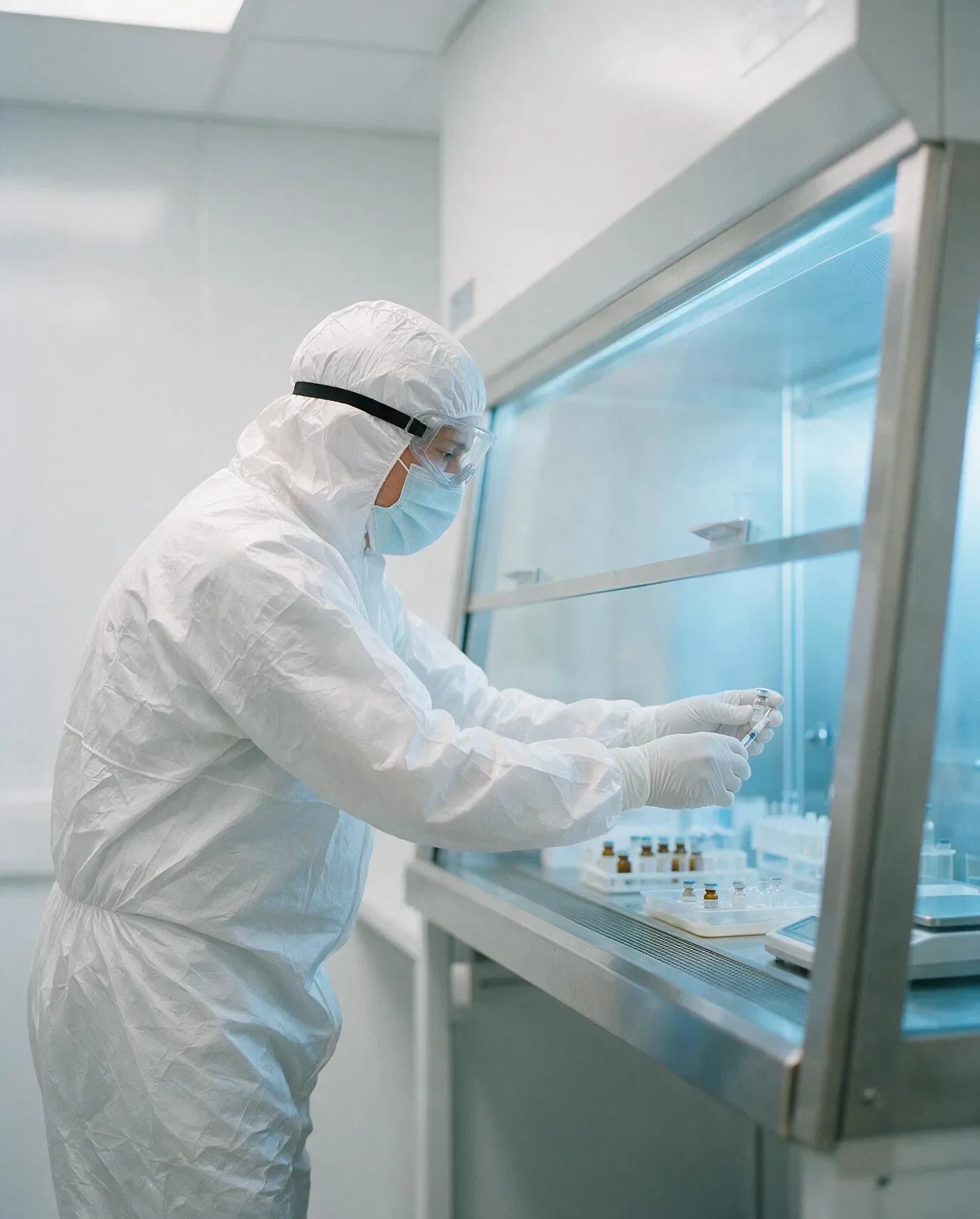 A technician in a white sterile gown, hood, goggles and mask holds a small vial inside a laminar airflow hood lined with vials